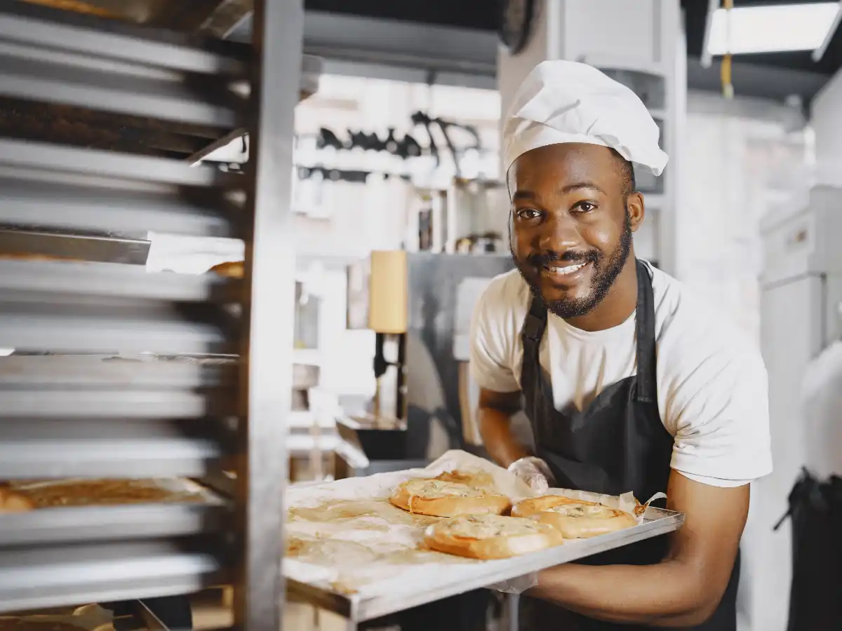 Smiling baker in a white hat and apron holds a tray of freshly baked pastries in an industrial kitchen. The atmosphere is warm and inviting.
