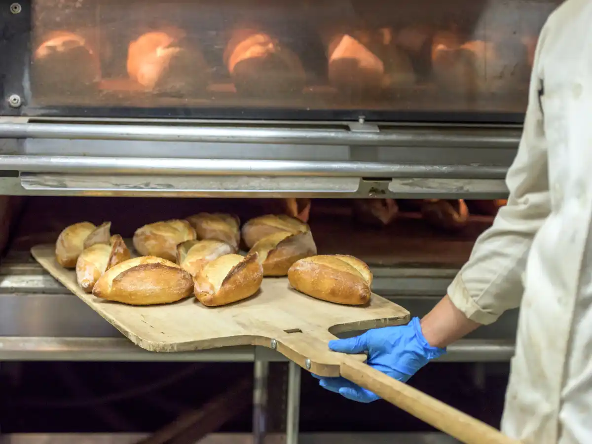 Baker in a white coat and blue glove uses a wooden peel to remove several golden brown loaves of bread from an industrial oven, conveying warmth and craftsmanship.