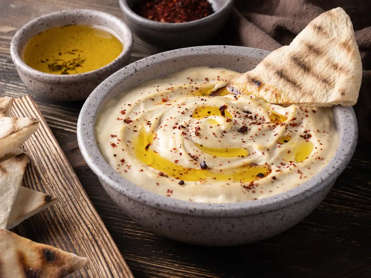 A bowl of creamy hummus topped with olive oil and spices, garnished with a pita slice. Olive oil and spice bowls are in the background on a wooden table.
