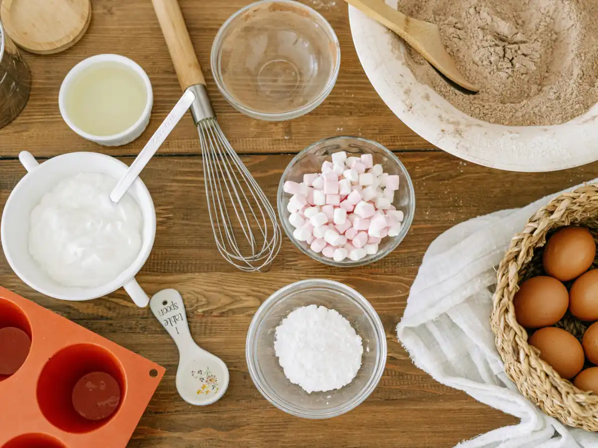 Baking ingredients on a wooden table: yogurt, oil, marshmallows, flour, whisk, eggs in a basket, measuring spoon, silicone mold, conveying a cozy, homemade vibe.