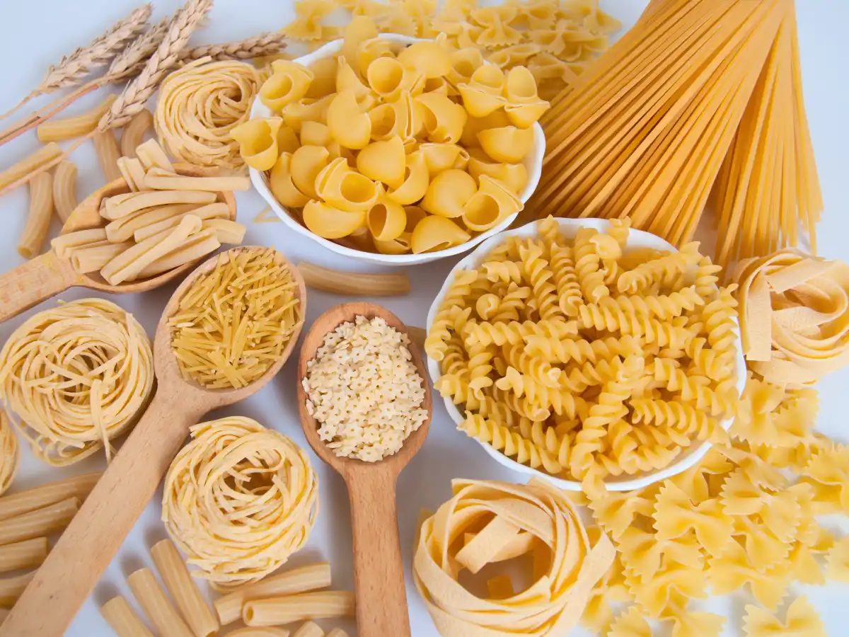 An assortment of uncooked pasta is displayed, including fusilli, spaghetti, and shells. Wooden spoons and bowls add a rustic touch, evoking a cozy, homely feel.