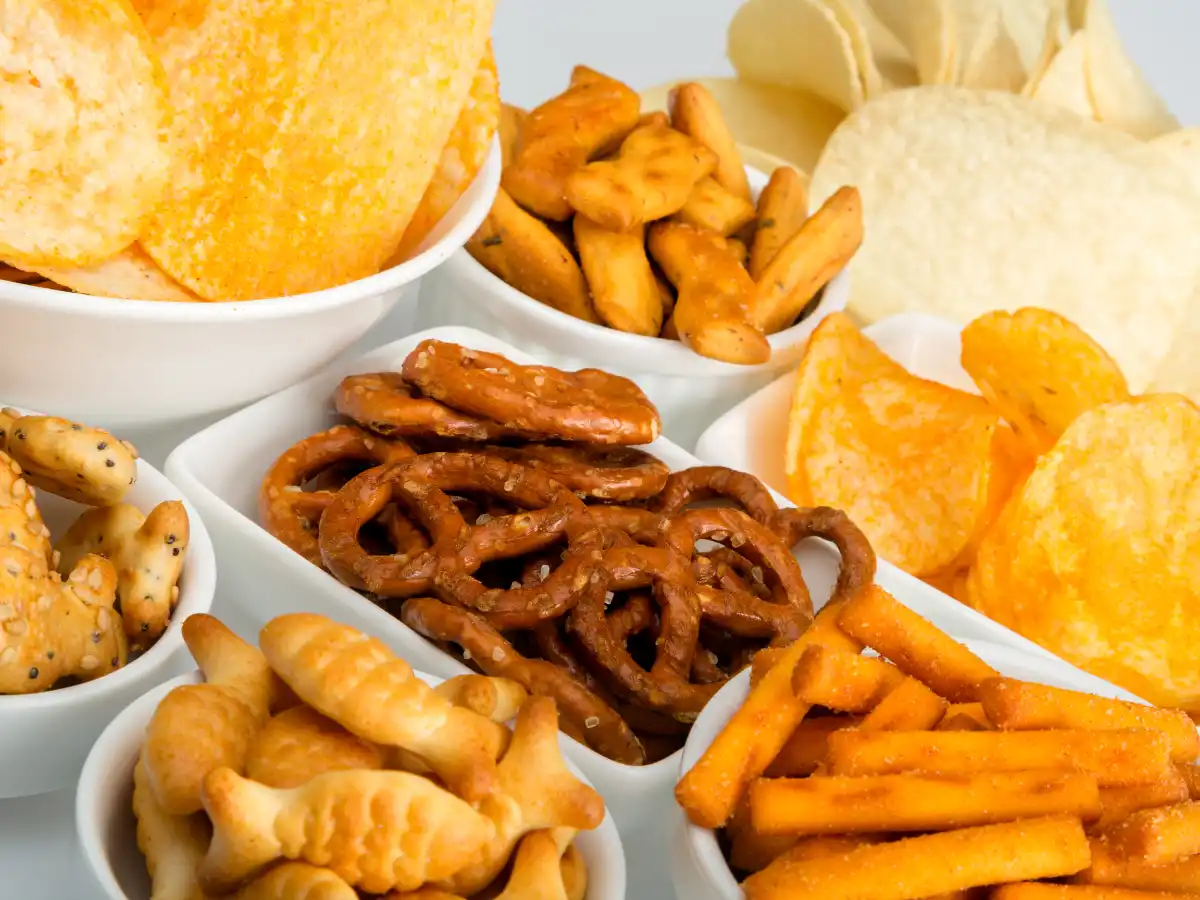 An assortment of snacks in white bowls, including pretzels, potato chips, cheeseballs, and breadsticks, creates a casual and inviting atmosphere.