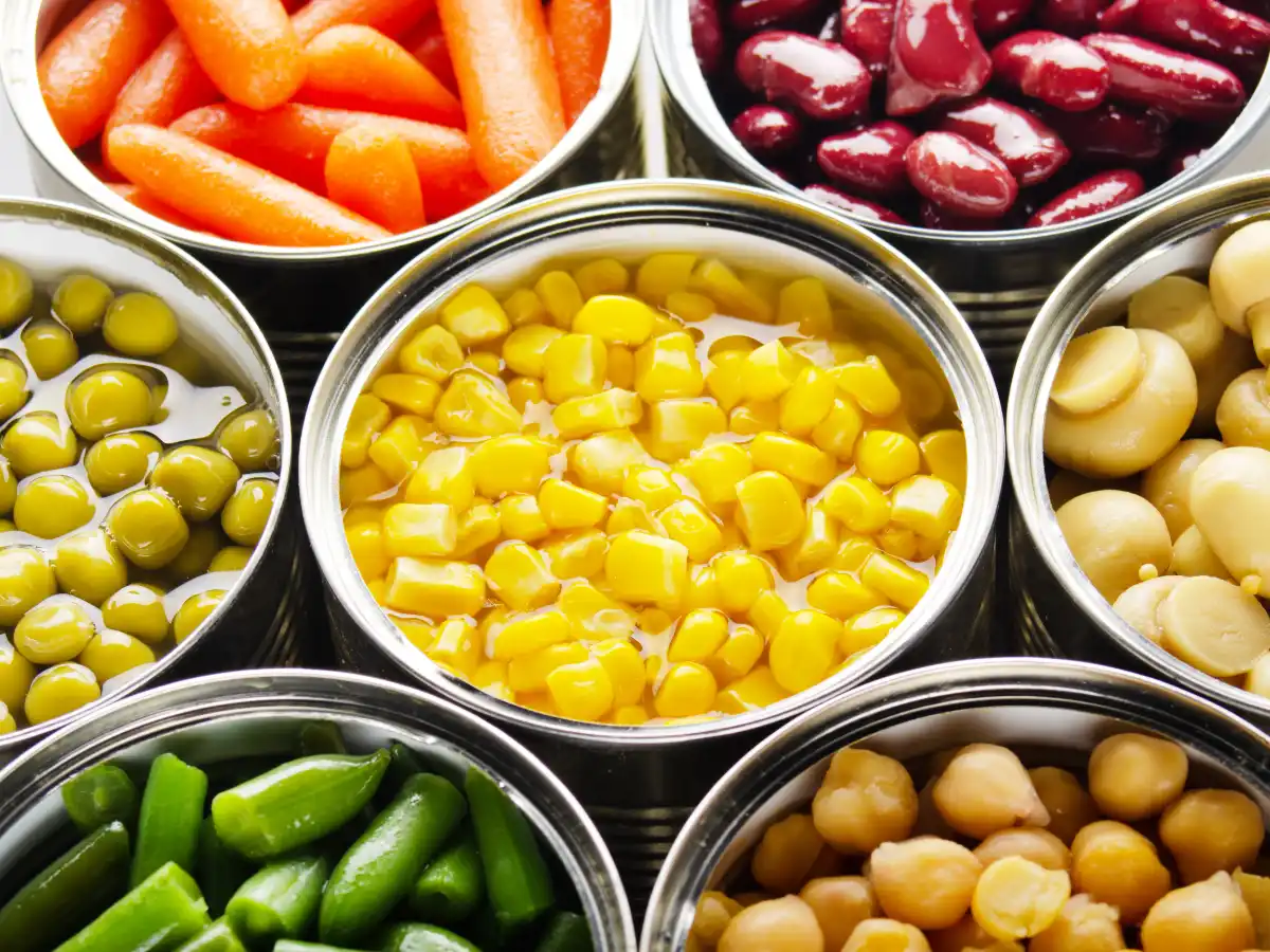 Close-up of open cans filled with colourful vegetables: bright carrots, red kidney beans, peas, corn, mushrooms, green beans, and chickpeas.