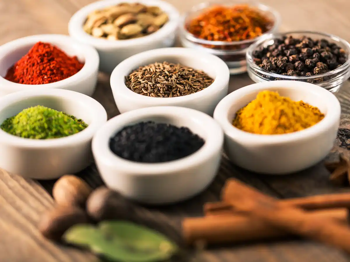 A variety of colourful spices in small white bowls on a wooden table. Visible spices include red, yellow, and black, conveying a vibrant culinary theme.
