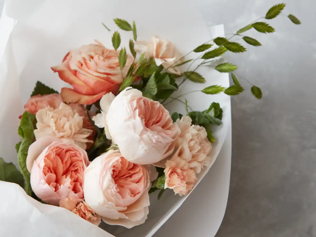 A bouquet of peach and pink roses and carnations wrapped in white paper with greenery. The arrangement is elegant and conveys a gentle, romantic mood.