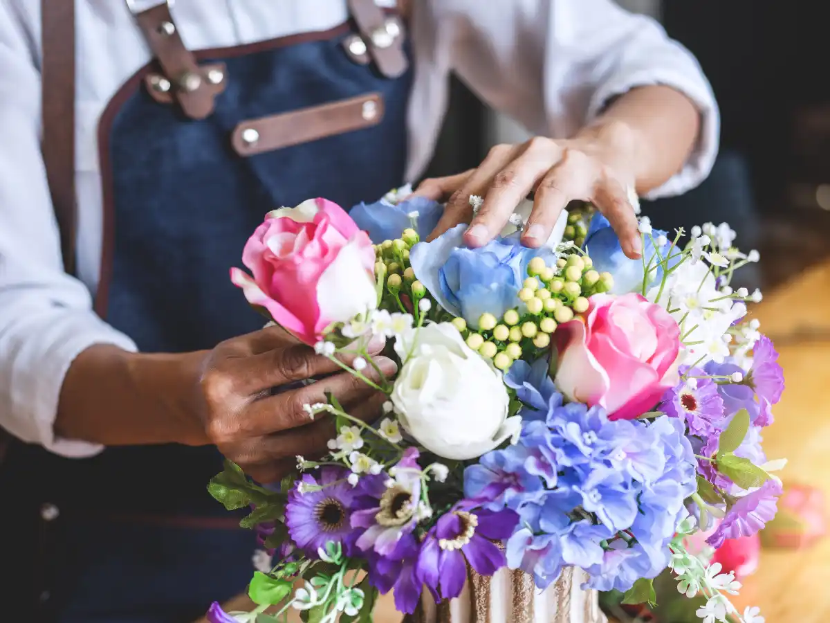 A florist arranges a vibrant bouquet of pink roses, white tulips, blue hydrangeas, and purple daisies. The scene conveys creativity and concentration.