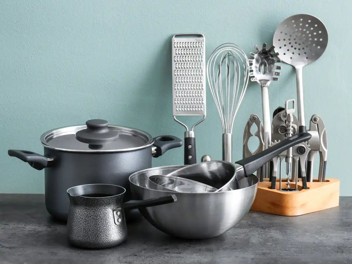 A collection of kitchen tools on a gray surface against a light green wall, including pots, a grater, whisk, bowls, and a utensil holder, creating a tidy and organized cooking setup.