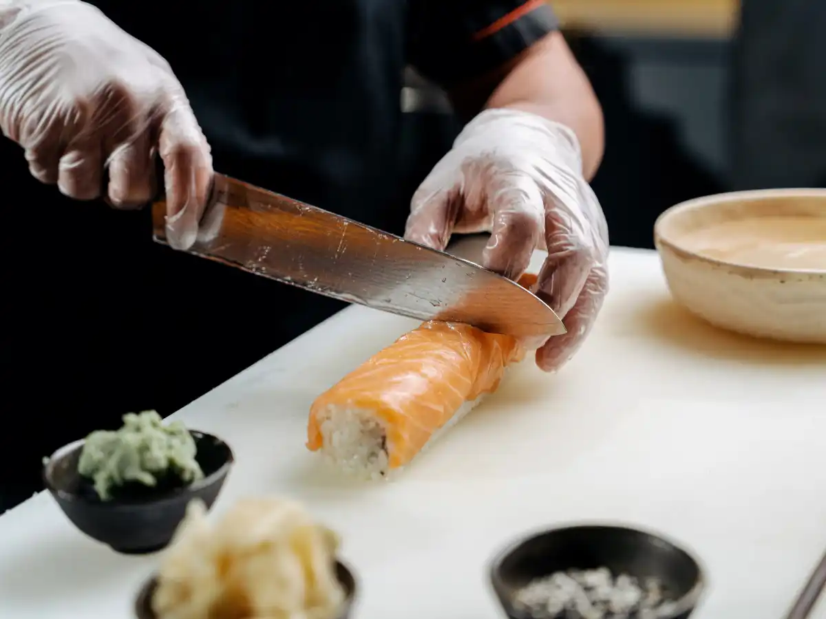 A sushi chef wearing gloves slices a salmon roll on a white cutting board. Bowls of wasabi, ginger, and sesame seeds are nearby, creating a focused culinary scene.