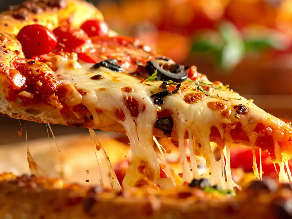 A slice of pizza is being lifted, showing melted, stretchy cheese. The pizza is topped with cherry tomatoes, black olives, and herbs, creating a tempting, savory look.