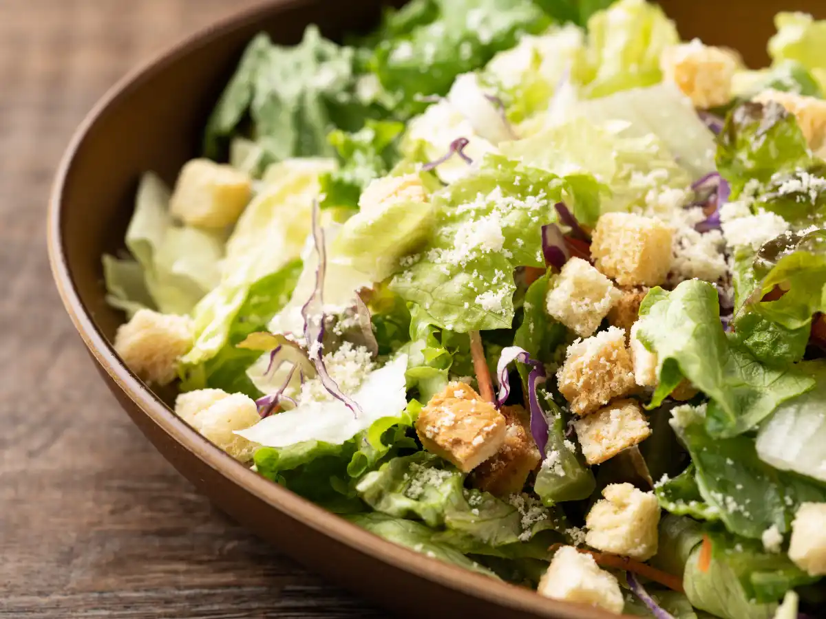 Close-up of a fresh Caesar salad in a brown bowl, featuring crisp lettuce, golden croutons, shredded cheese, and purple cabbage on a wooden table.