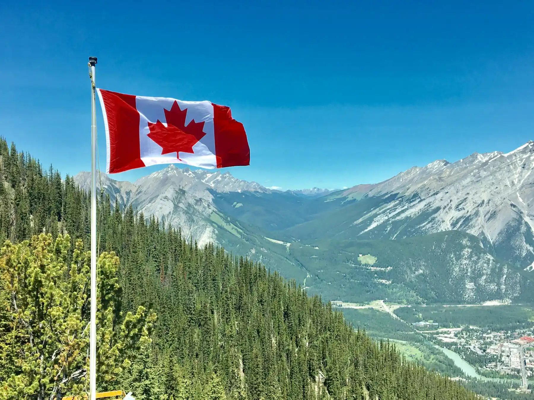 Canadian flag waving above a lush green forest beneath clear blue skies, with majestic mountain ranges in the background. The scene conveys tranquility and natural beauty.