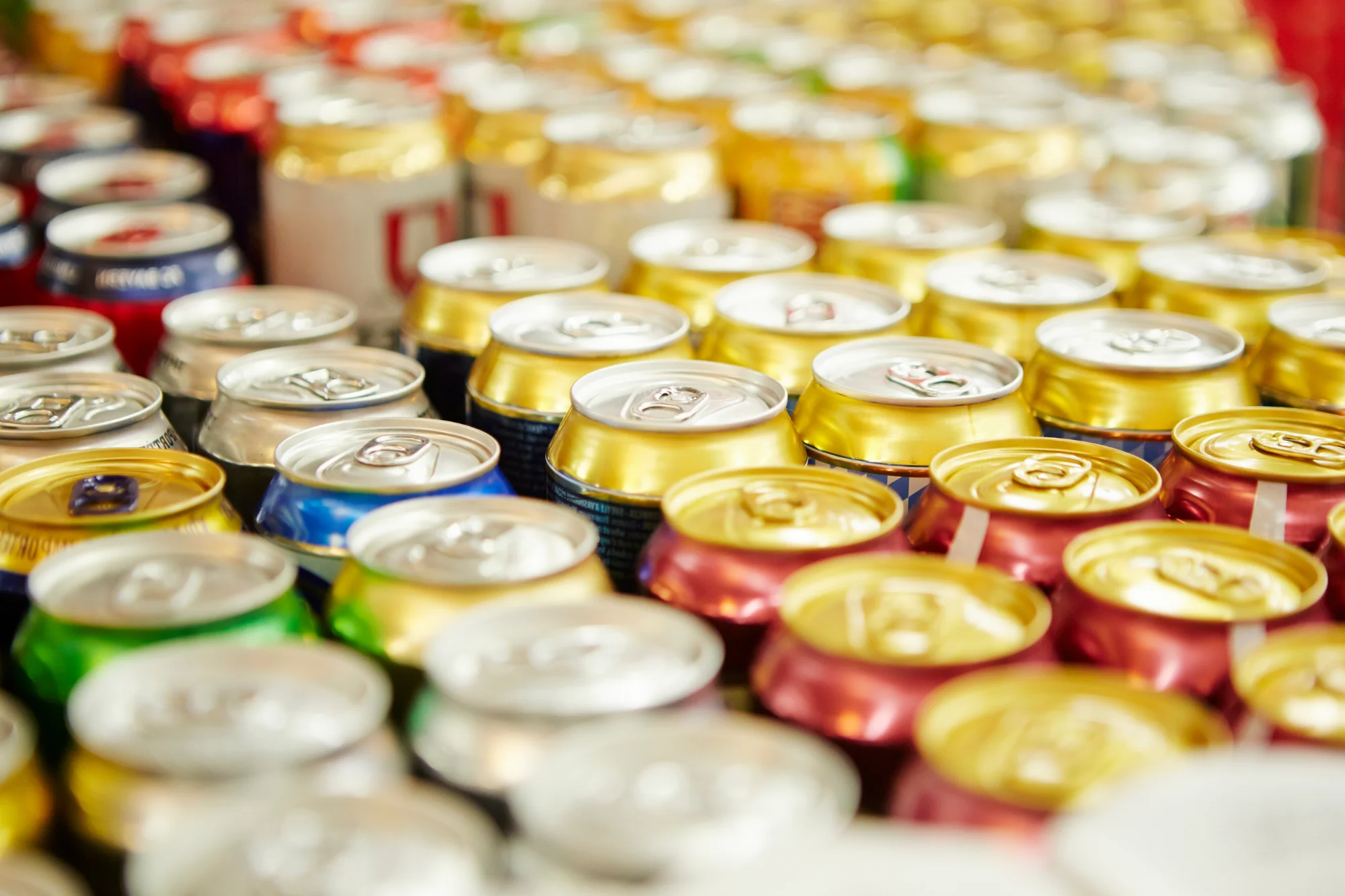 Rows of assorted colorful beverage cans tightly packed together, with many silver, gold, red, green, and blue tops visible, suggesting a variety of drinks like sodas or beers.
