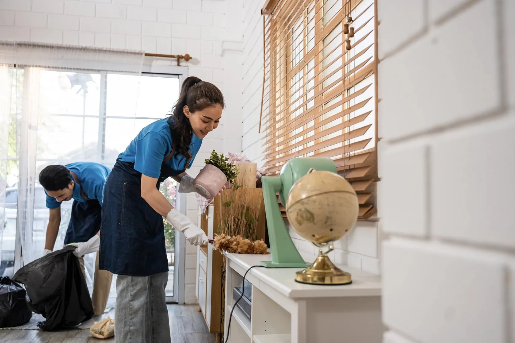 Two cleaners in matching aprons tidying a bright home, arranging a potted plant and decor.