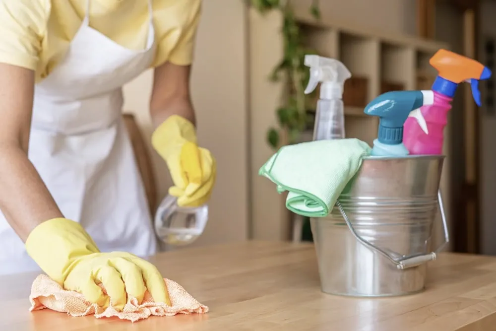 Cleaner in yellow rubber gloves wiping down a kitchen counter beside a bucket of cleaning supplies.