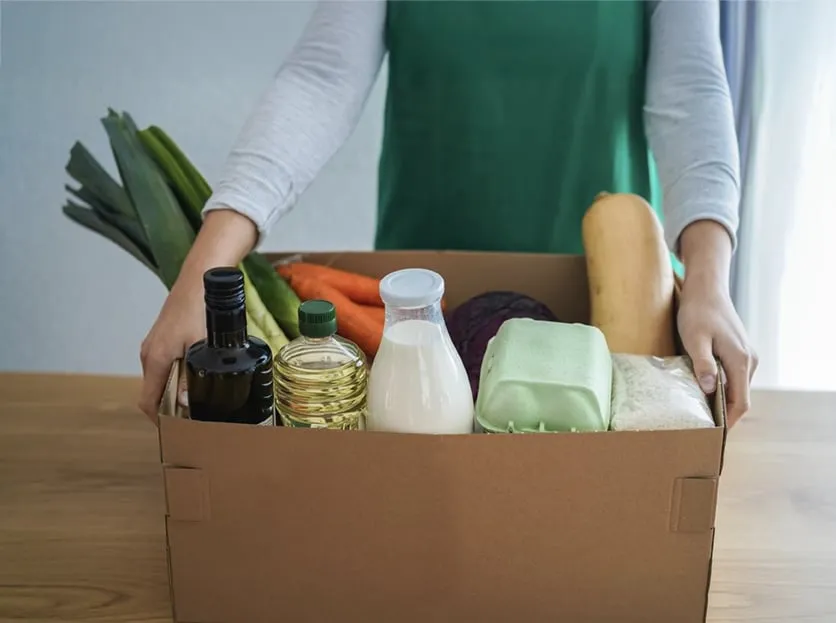 Person in a green apron packing a wooden box with fresh groceries.
