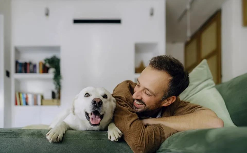 Man relaxing on a couch, laughing while cuddling a happy white dog.