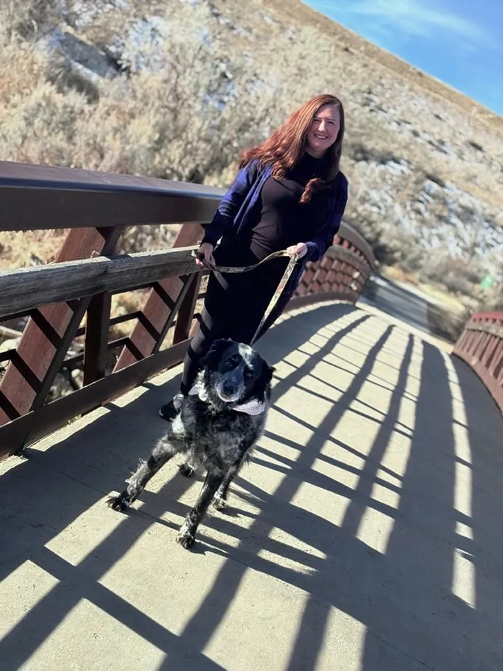 Smiling woman walking a leashed dog across a bridge with mountain scenery in the background.