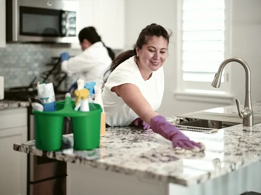 Cleaner in rubber gloves wiping down a granite kitchen countertop while a colleague cleans nearby.