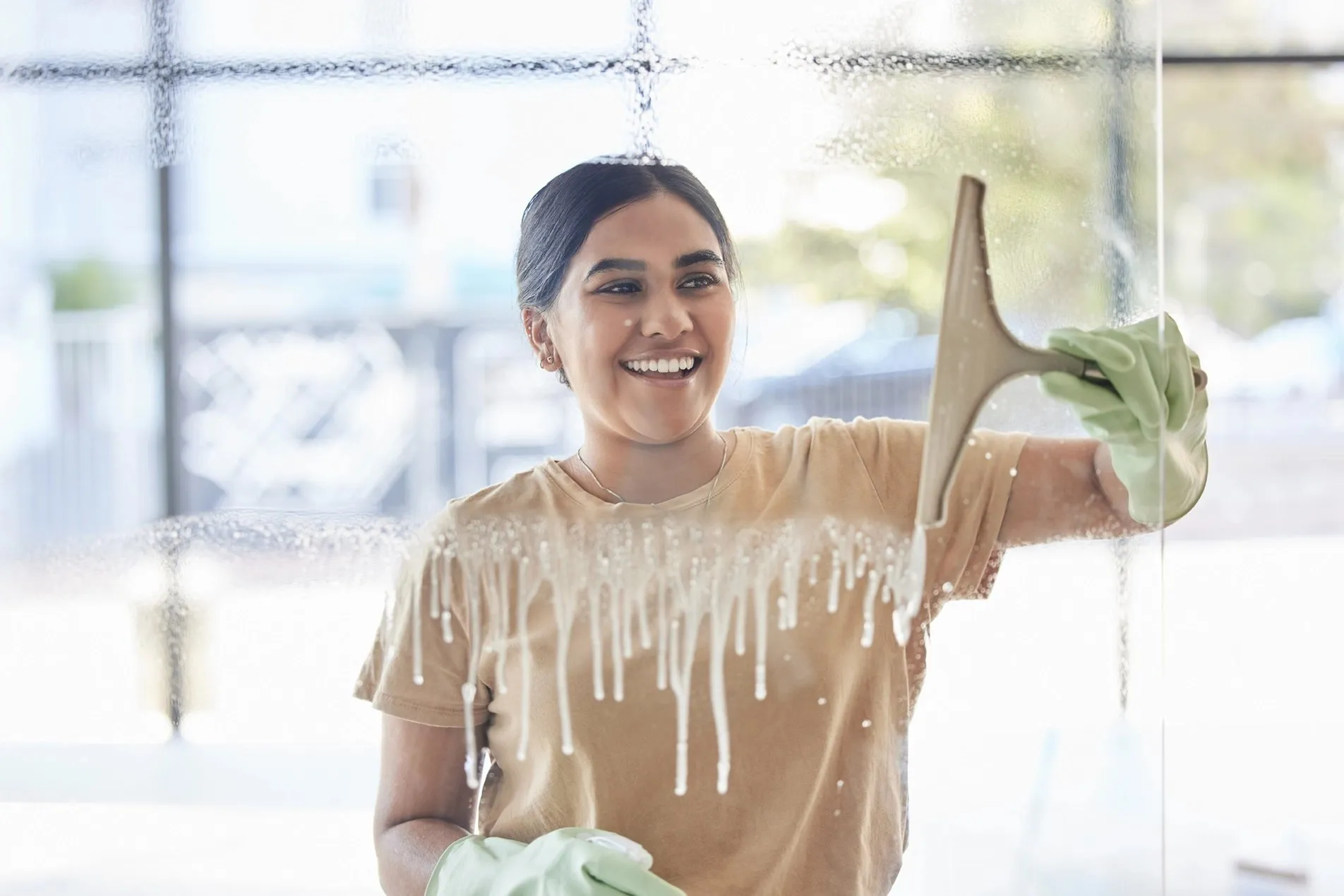 Smiling woman using a squeegee to clean soap suds off a large window pane.