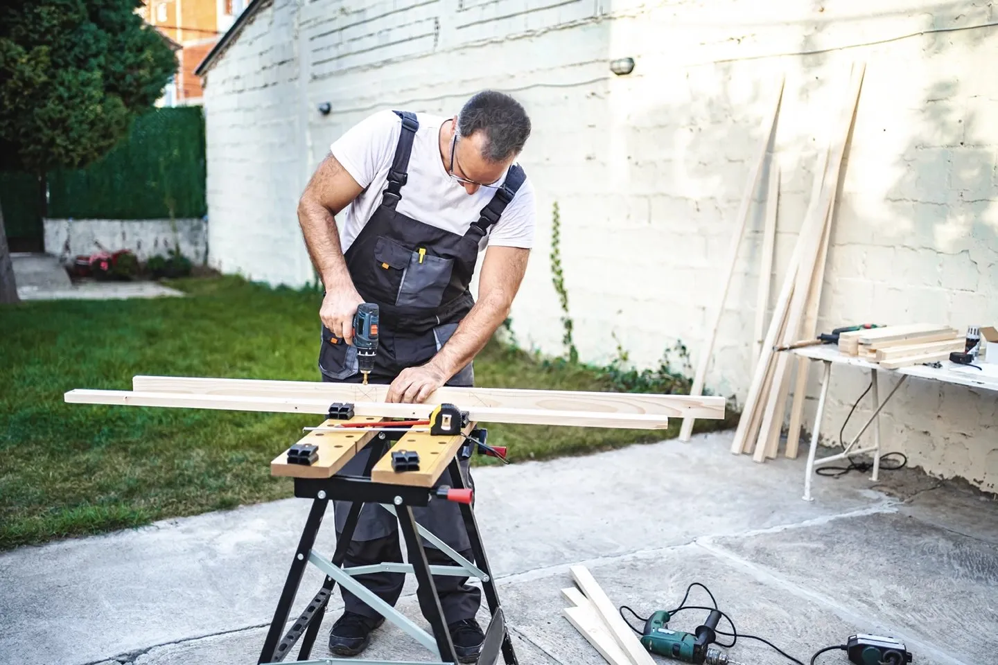 Handyman using a power drill on a wood plank secured in a clamp in a backyard.