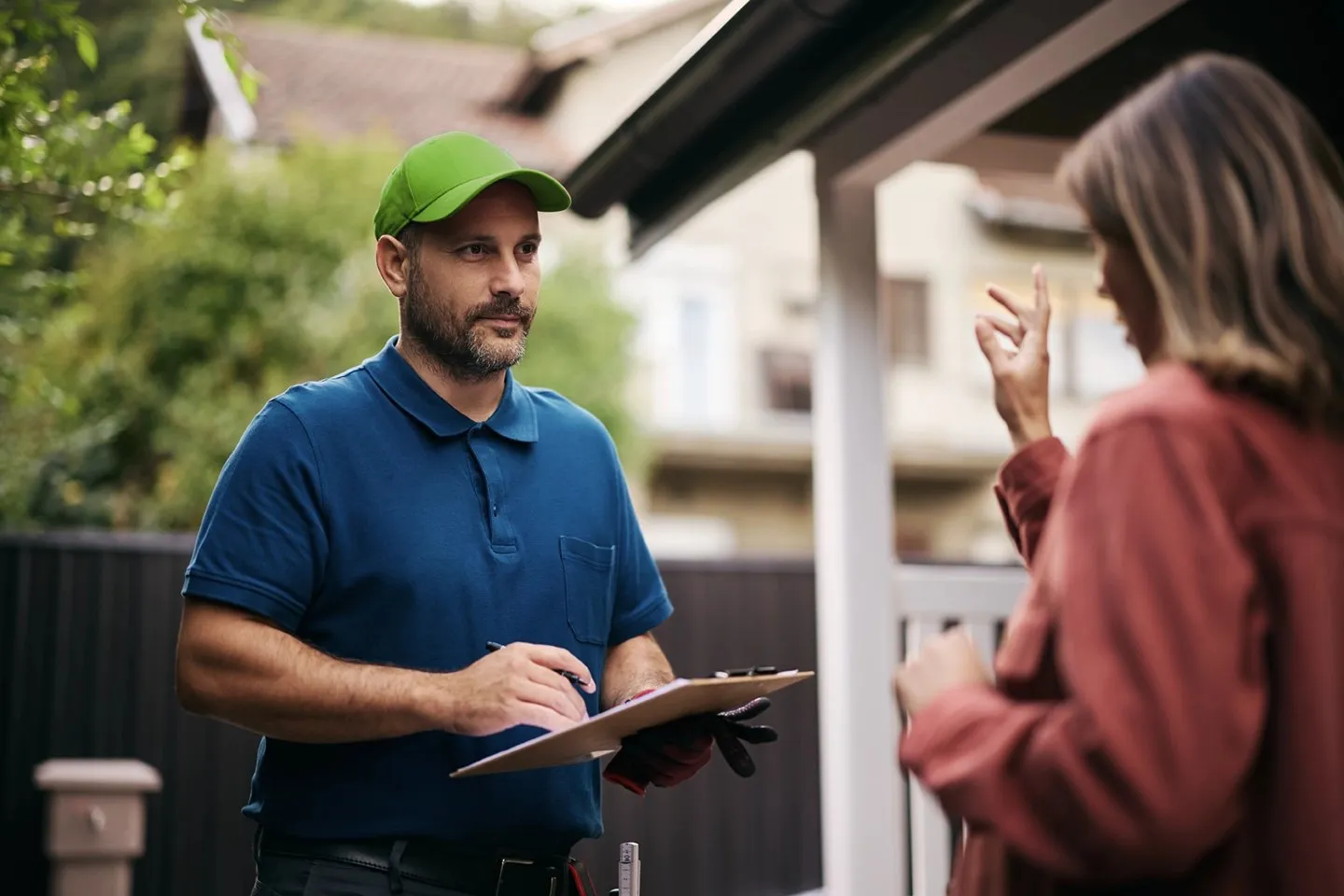 A property manager in a green cap reviewing details with a homeowner outside her house, clipboard in hand.