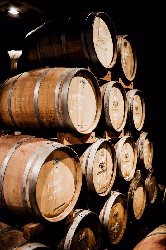 Stacked wooden wine barrels in a dimly lit cellar, with labels indicating various types and years.