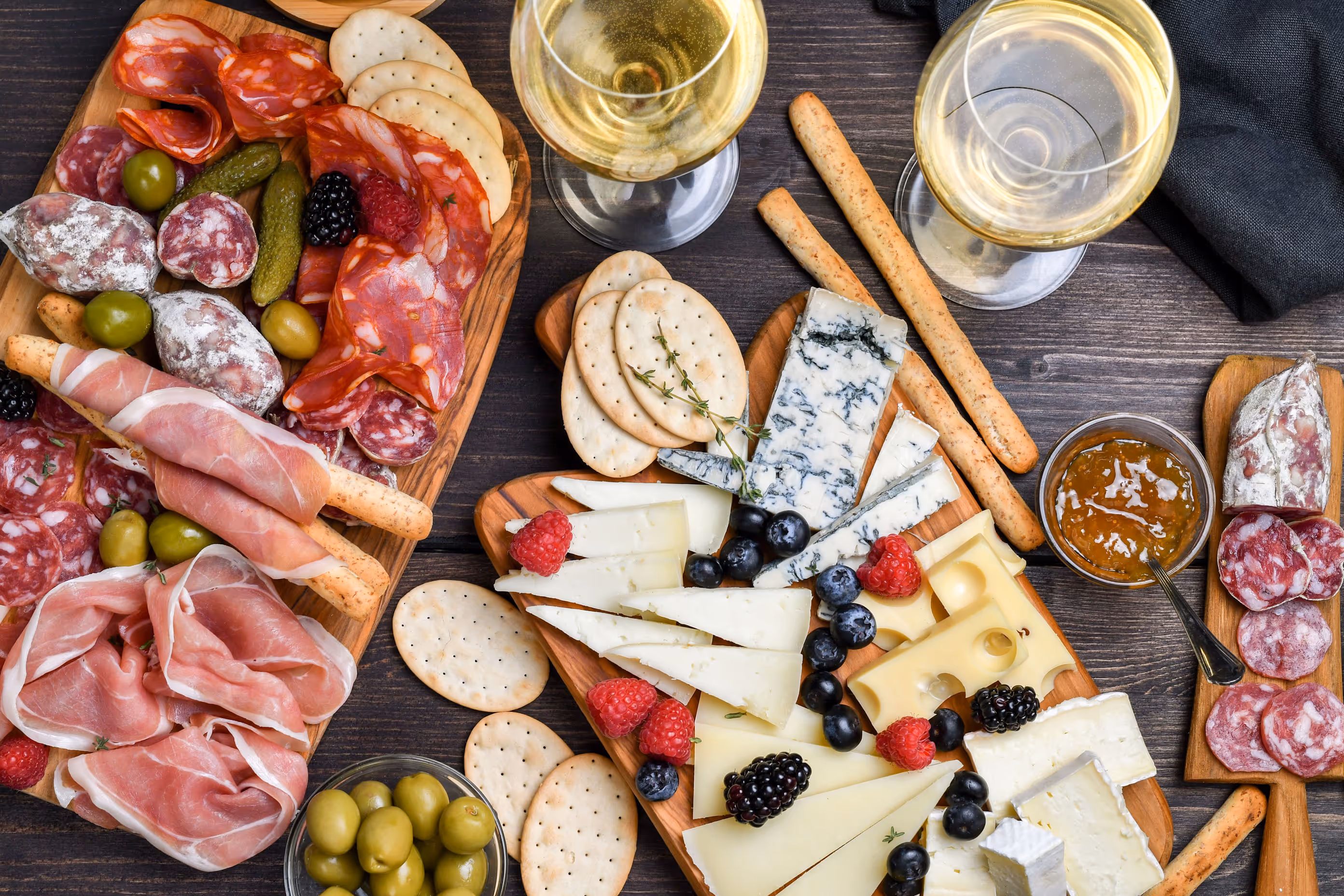 Charcuterie boards with assorted cheeses, meats, olives, crackers, and berries. Two glasses of white wine, breadsticks, and a bowl of marmalade add elegance.