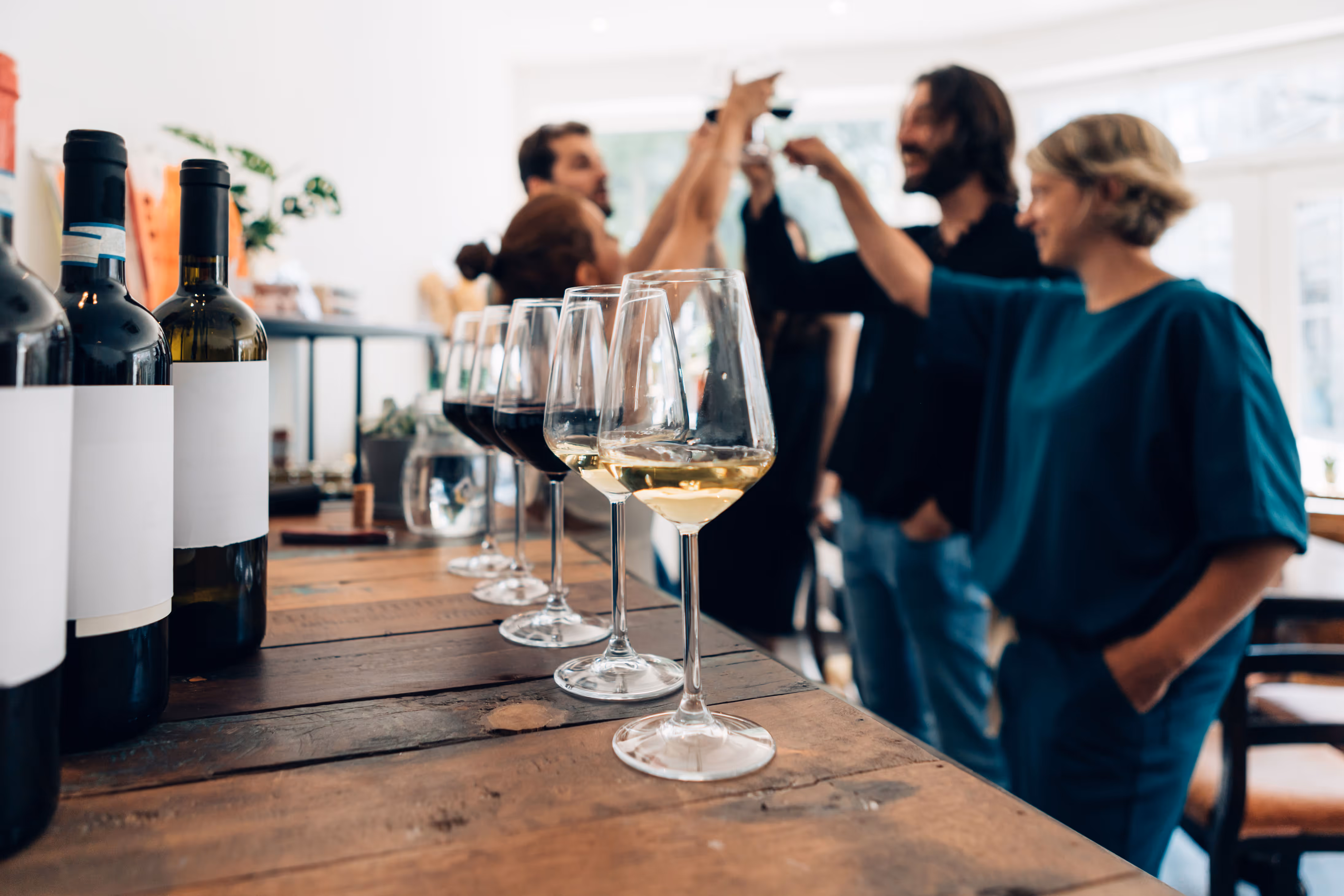 A row of red and white wine glasses with bottles on a wooden table, while a group of people in the background raise glasses in a joyful toast.