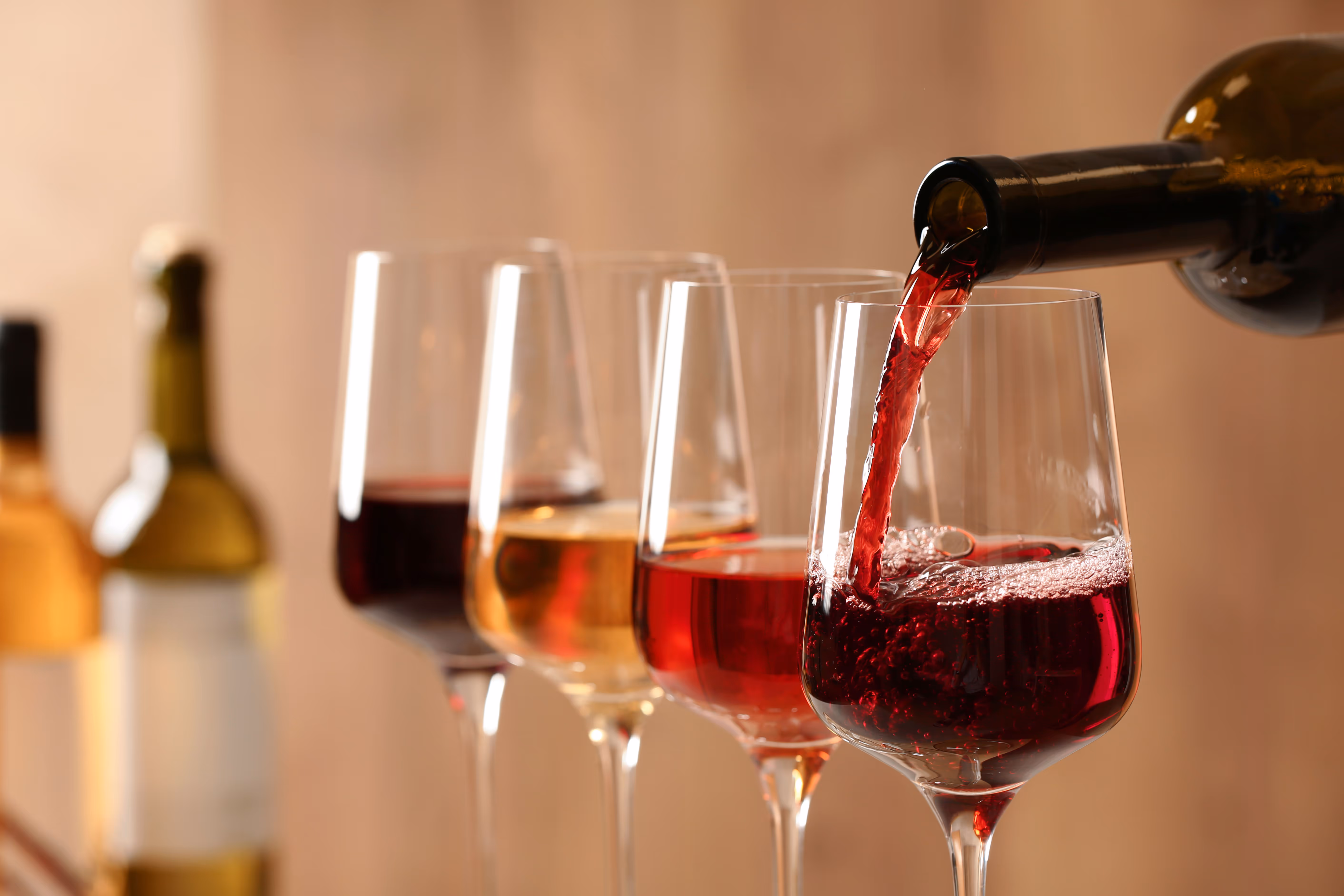 Four glasses of wine are arranged in a row. A bottle is pouring red wine into the nearest glass. The background is softly blurred, creating a warm, elegant atmosphere.