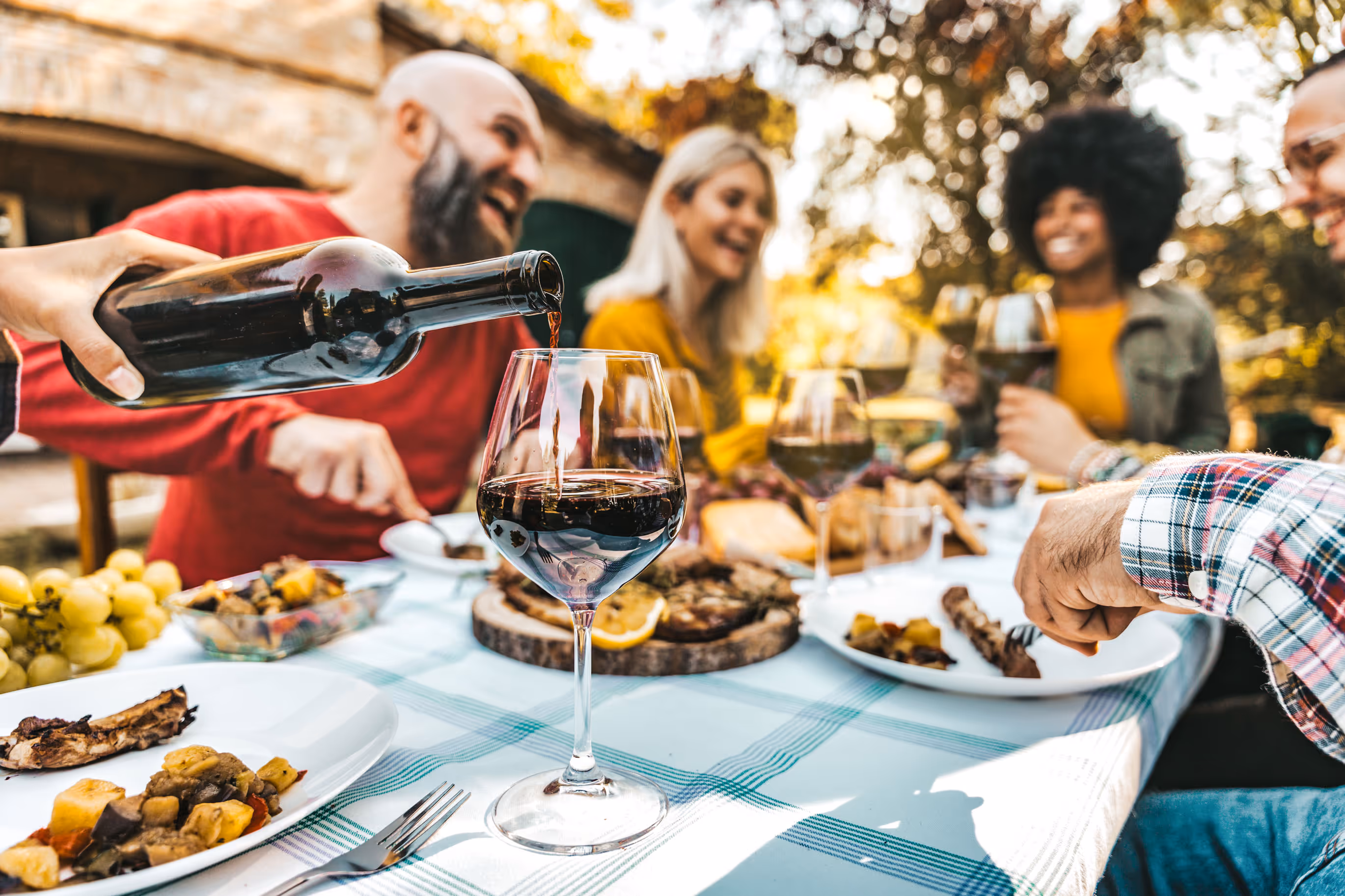 Five friends enjoy an outdoor meal, smiling and chatting. A hand pours red wine into a glass. Plates of food and grapes are on the table.
