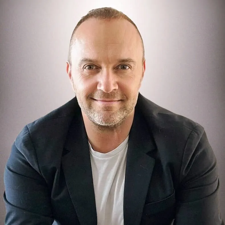 Smiling bald man with light stubble wearing a white shirt and black blazer against a plain background.