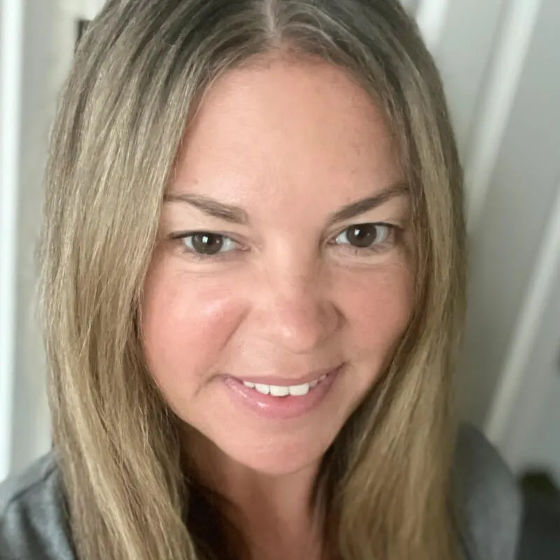 Close-up of a smiling woman with long blonde hair and brown eyes wearing a gray top.