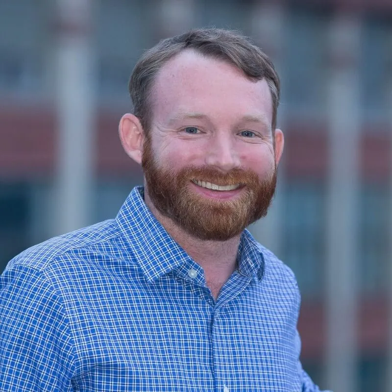 Smiling man with red beard and blue checkered shirt standing outdoors with blurred background.
