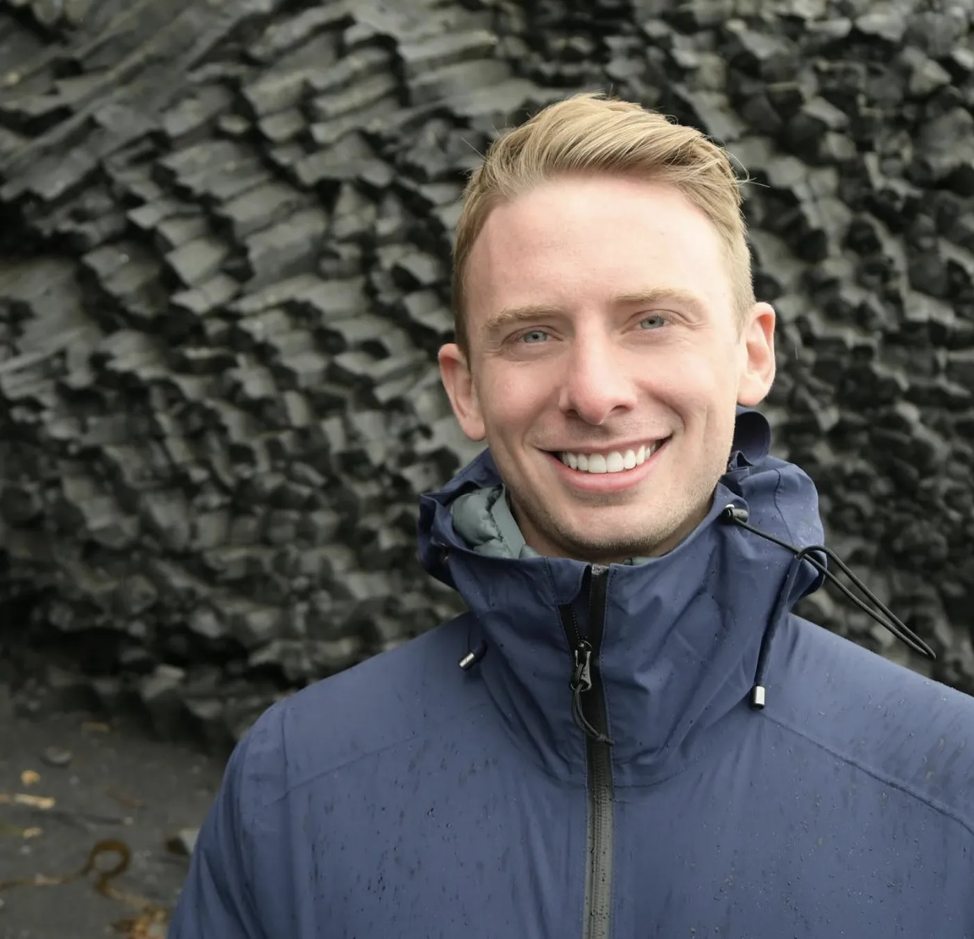 Smiling man with short blond hair wearing a blue jacket standing in front of hexagonal basalt rock formations.