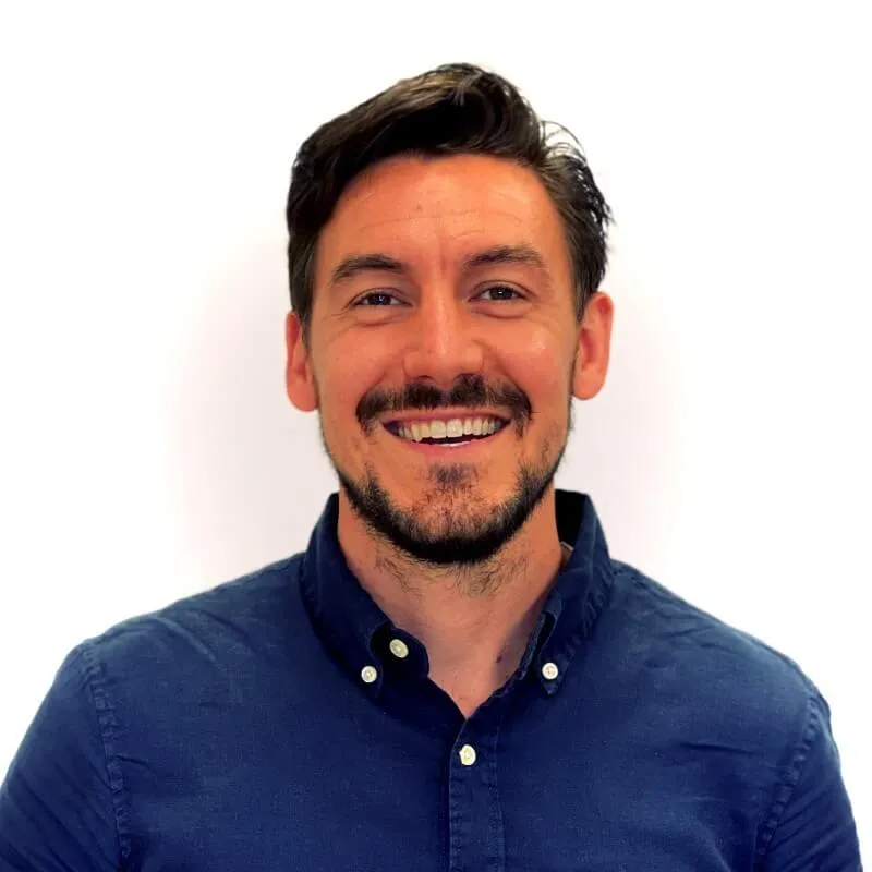Smiling man with dark hair and beard wearing a navy blue button-up shirt against a white background.