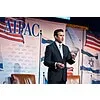 Man in a suit speaking at a podium with American flags and a backdrop featuring the AIPAC logo.