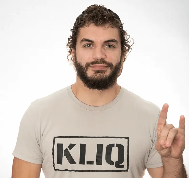 Man with curly hair and beard wearing a beige KLJQ t-shirt, making a hand sign with his right hand.