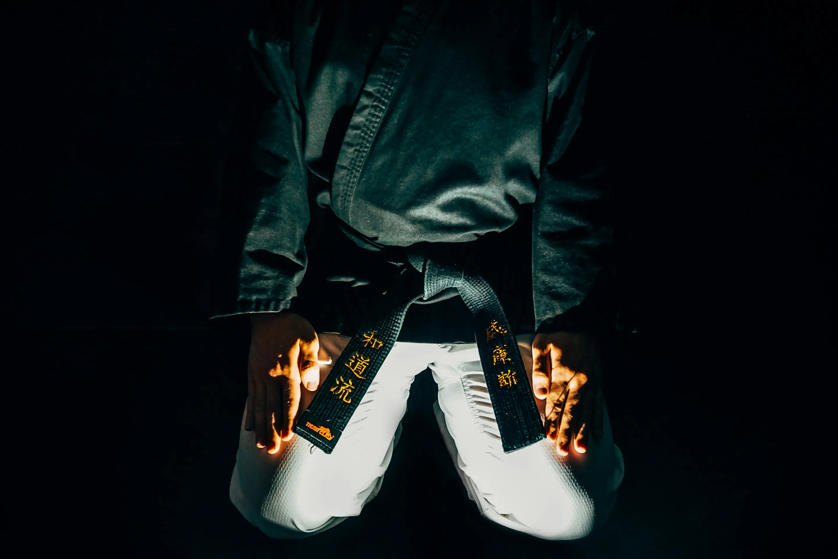 Person kneeling wearing a black martial arts gi with a black belt and white pants, hands resting on thighs.