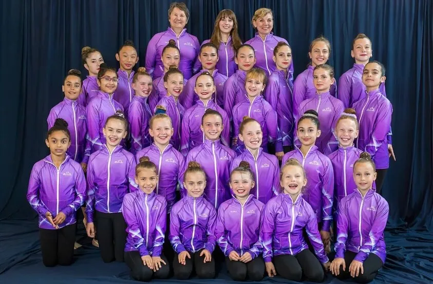 Large group of young female gymnasts and three coaches posing in matching purple jackets against a dark backdrop.