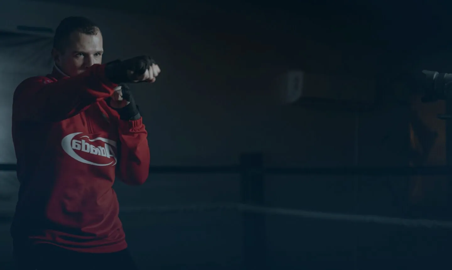 Man in a red sweatshirt and black gloves throwing a punch inside a dimly lit boxing ring.