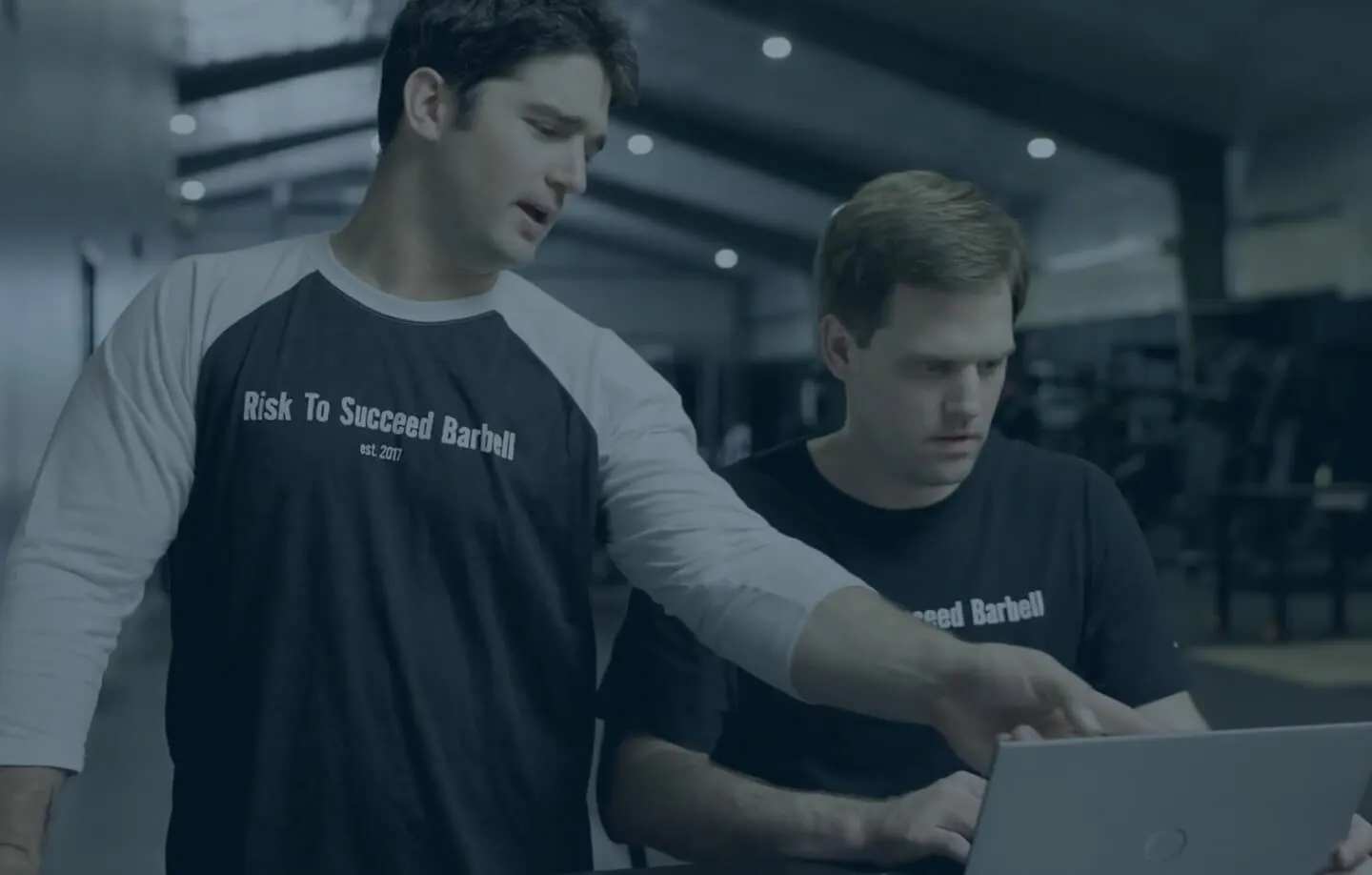 Two men in a gym, one pointing at a laptop screen while the other looks at it attentively, both wearing 'Risk To Succeed Barbell' shirts.