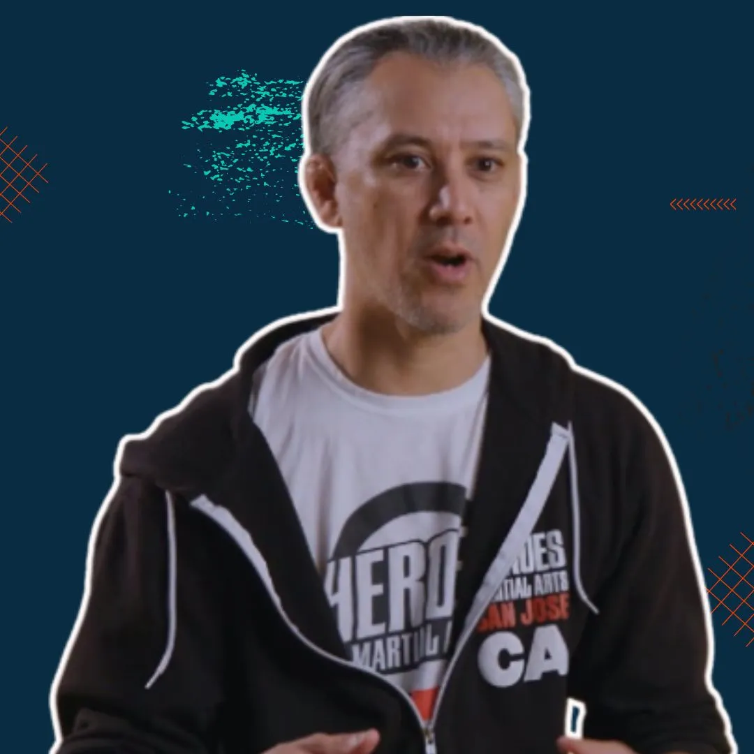 Man with short gray hair wearing a black hoodie over a white martial arts t-shirt, speaking with a dark blue background.