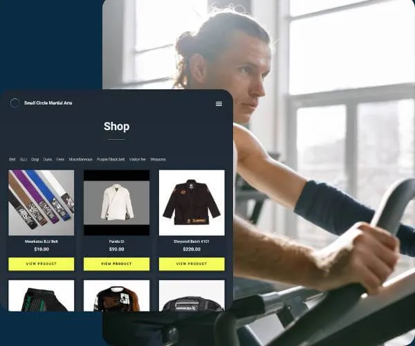 Man with tied-back hair wearing a black arm sleeve, exercising on a stationary bike in a bright room with large windows.