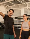 Man in black sportswear pointing while standing next to woman in gray sports bra and black leggings inside a gym.