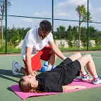 A personal trainer assisting a woman doing sit-ups on a pink mat on an outdoor tennis court.