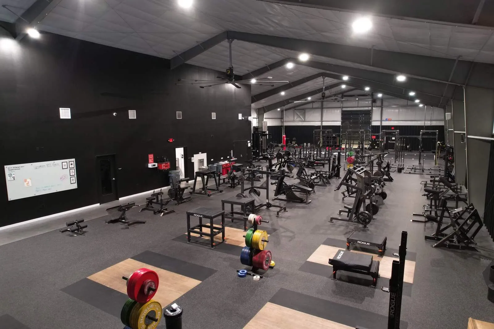 Spacious indoor gym with weightlifting platforms, racks of colorful weight plates, various strength training equipment, and black walls under bright ceiling lights.