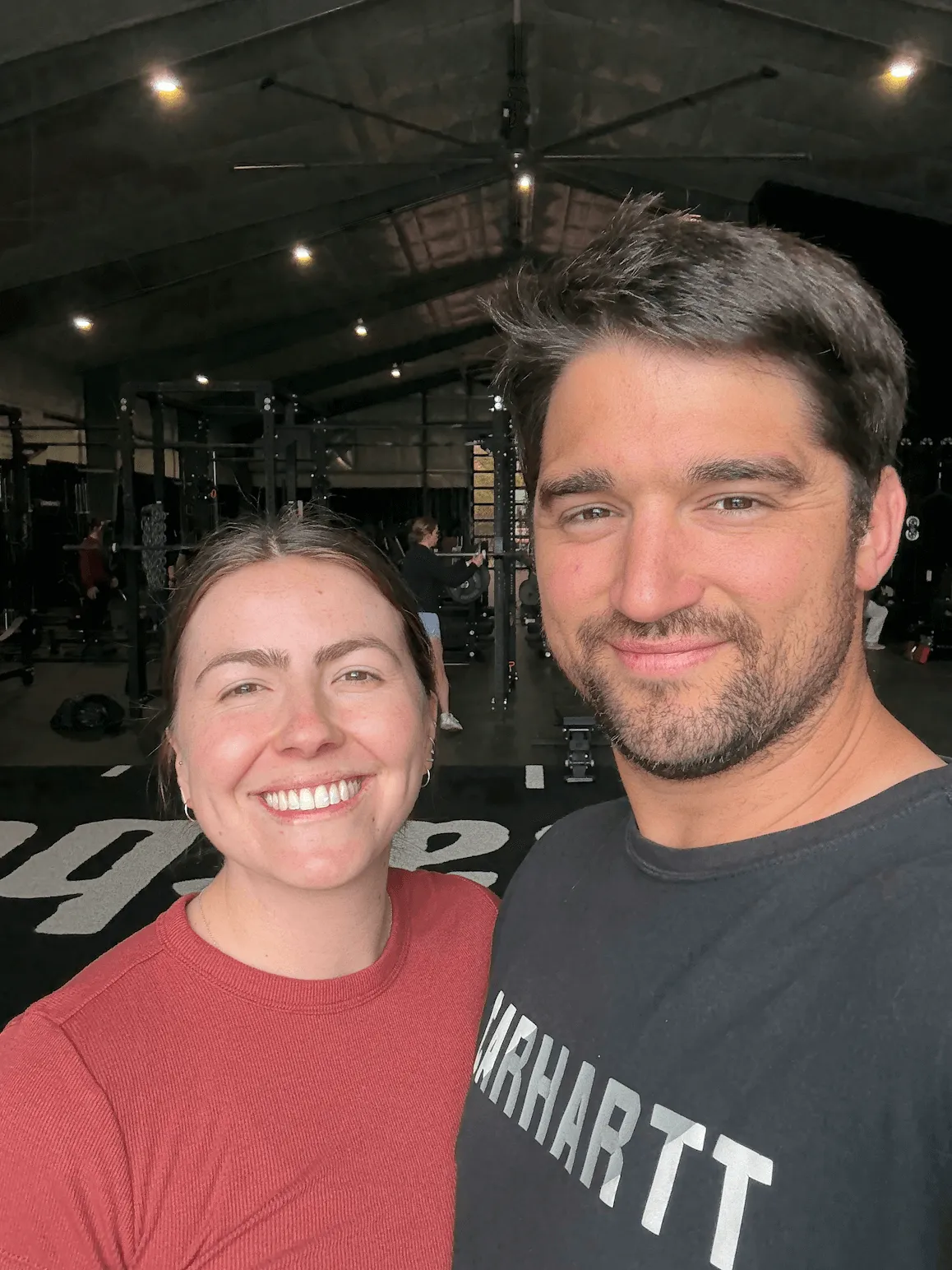 Smiling man and woman taking a selfie inside a gym with workout equipment in the background.