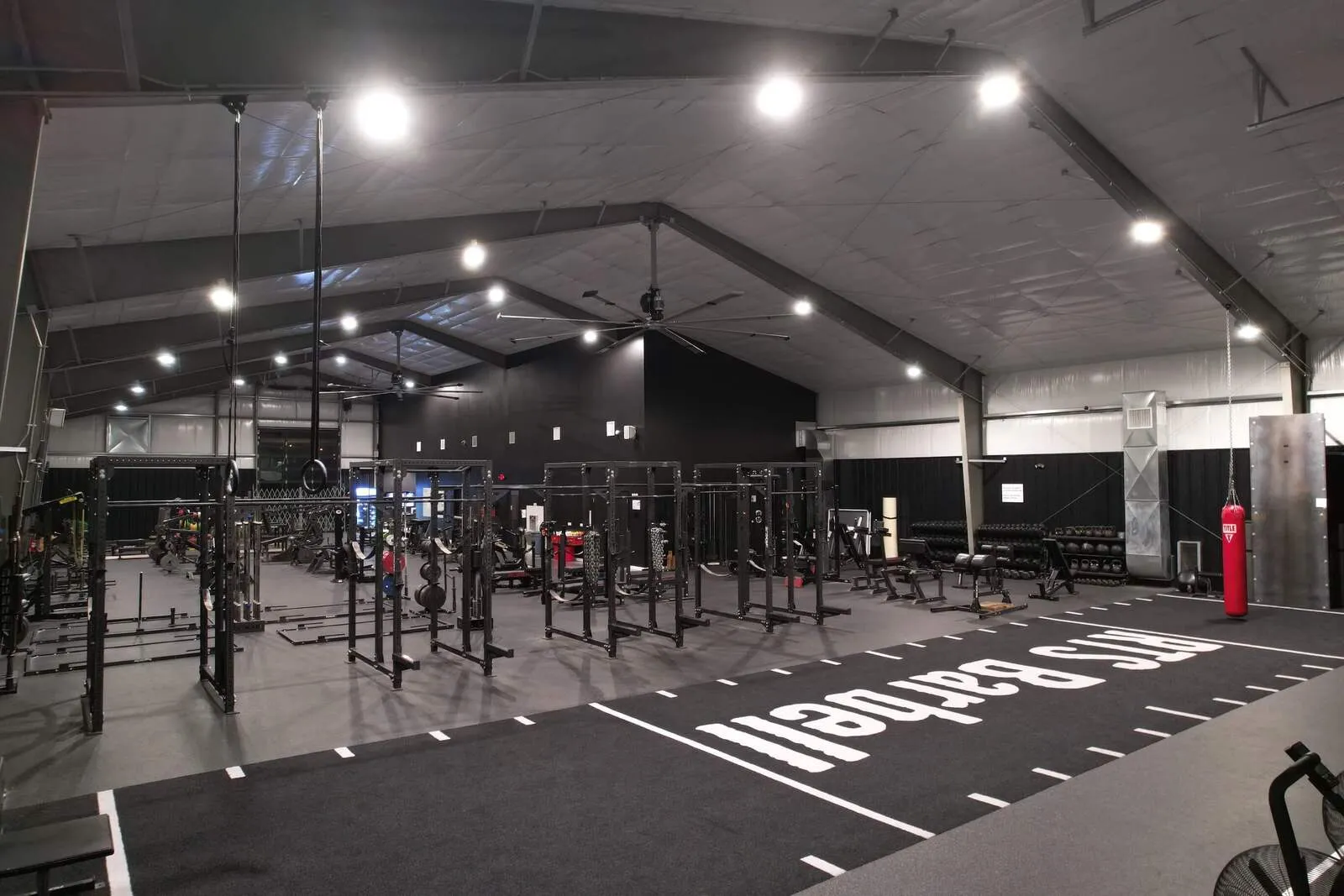 Spacious indoor gym with multiple weightlifting racks, gymnastic rings, punching bag, and workout equipment.