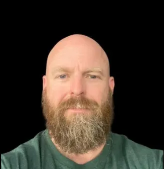 Close-up of a bald man with a full beard wearing a green shirt against a black background.
