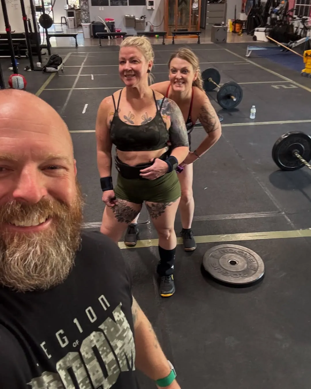 Three smiling people in workout attire posing for a selfie inside a gym with weightlifting equipment.
